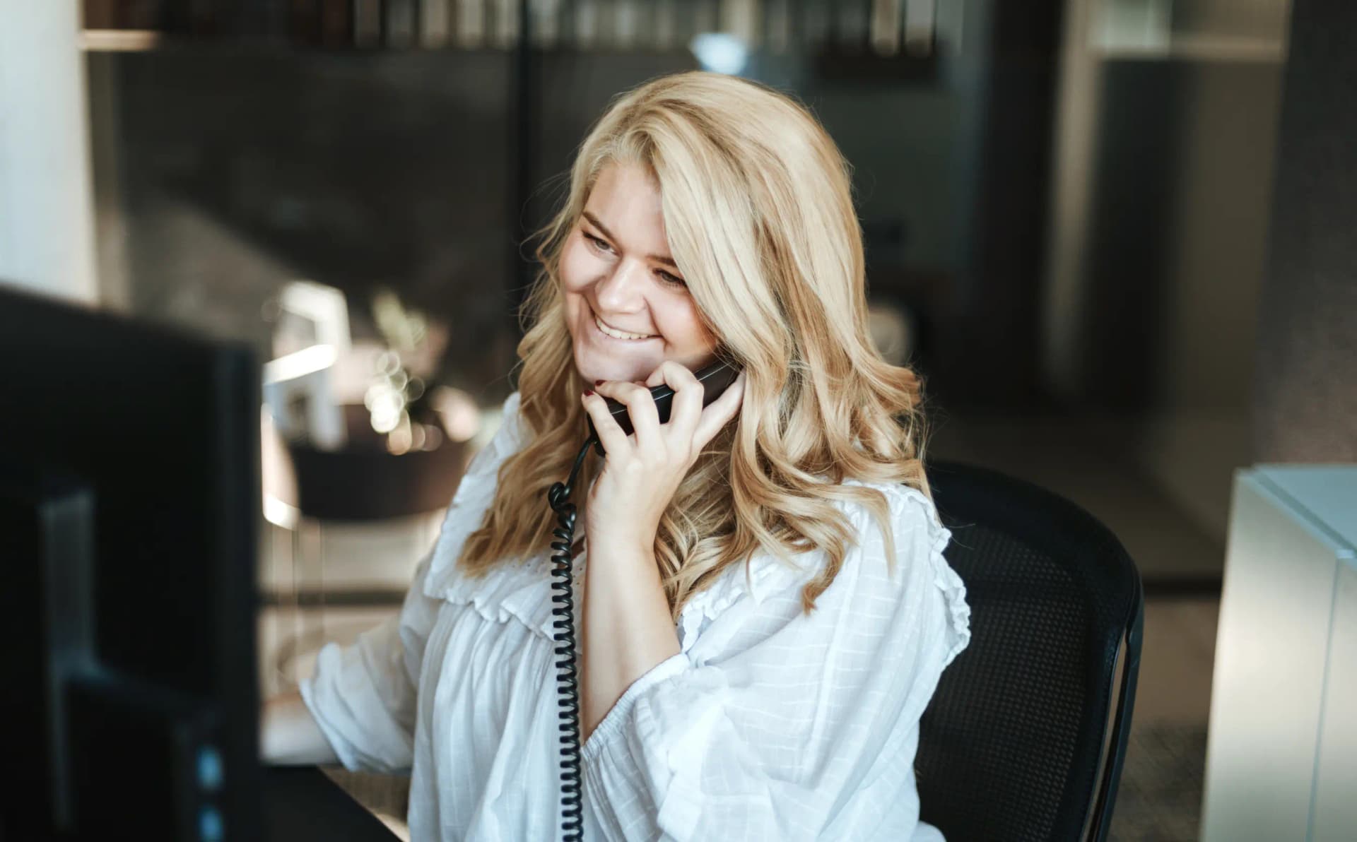 Eine lächelnde junge Frau mit blonden Haaren telefoniert an ihrem Arbeitsplatz im Büro.
