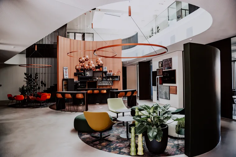 Modern lounge with colorful seating, a bar and stylish decor, lit by natural light from a large skylight.