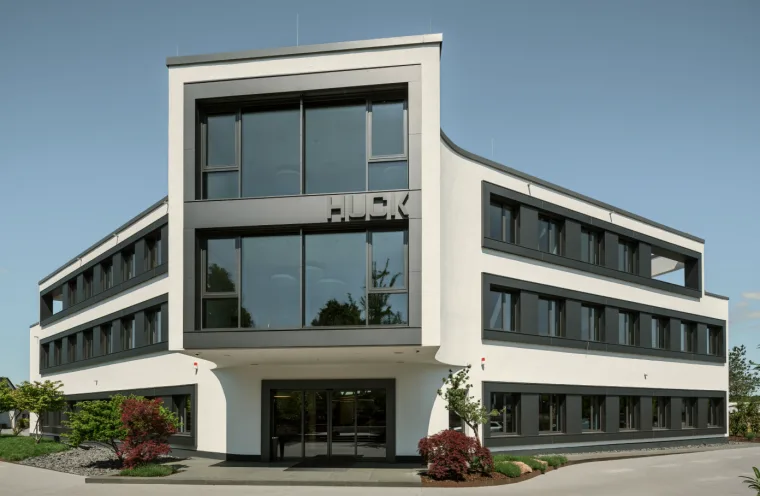 Modern office building with large glass windows and the lettering HUCK, surrounded by greenery and a clear sky.