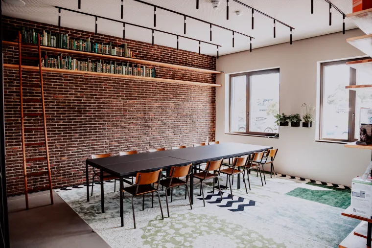 A spacious meeting room with a long table surrounded by wooden chairs, brick walls, large windows and a library ladder.