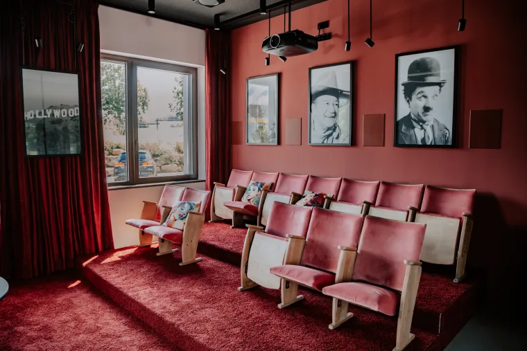 Cozy screening room with red walls, plush armchairs and classic black-and-white actor portraits, with a projector overhead.