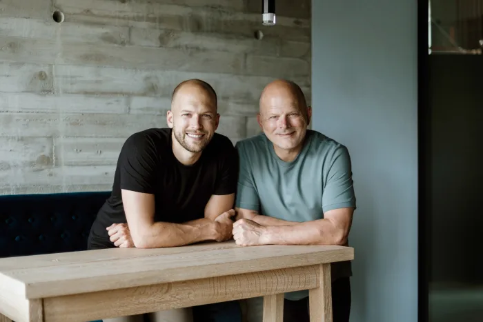 Two men sit at a wooden table in a modern, minimalist room with a concrete wall, leaning forward with a relaxed expression.
