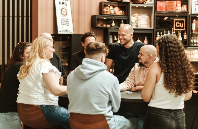 A group of people sitting around a table chatting, against the backdrop of a stylish bar with shelves of drinks.